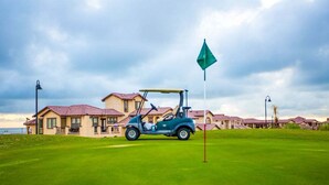 A golf cart on a green with a flag, a building with red roofs, and a cloudy sky.