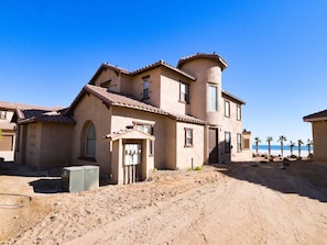 A two-story building with a flat roof, a small attached structure, and a sandy ground.