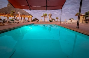 Ein Schwimmbecken mit Liegestühlen und Sonnenschirmen, umgeben von Palmen und einem strohgedeckten Gebäude.