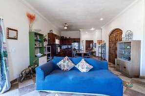A living room with a blue sofa, a green bookshelf, and a kitchen in the background.