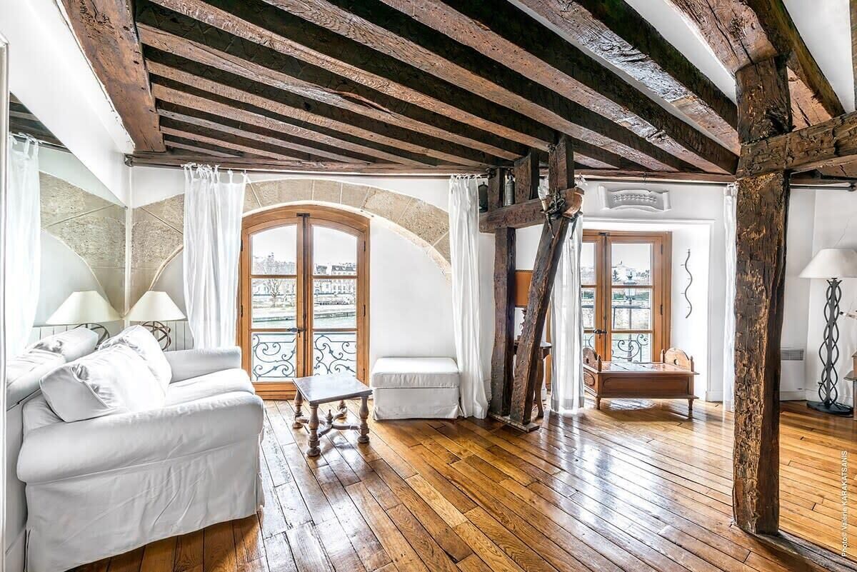 A room with a white sofa, a wooden bench, and a small table. There are two windows with white curtains, and a wooden beam ceiling.