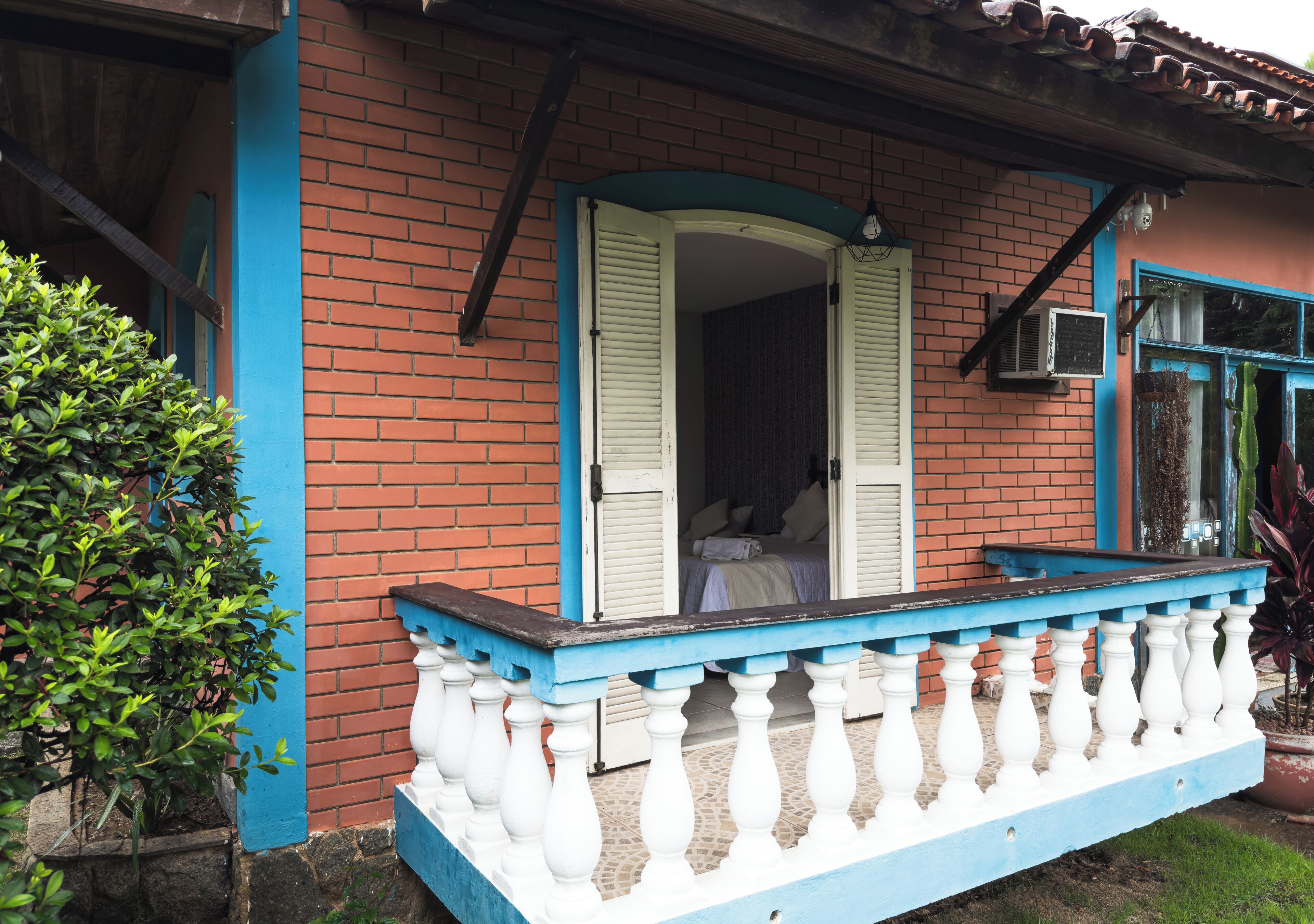 Un edificio de ladrillo con un balcón azul y blanco, una puerta con contraventanas y un pequeño equipo de aire acondicionado.