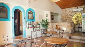 A dining area with round tables and chairs, a bookshelf, and a stone wall with a decorative window. - Pousada Flor De Penedo (Itatiaia)