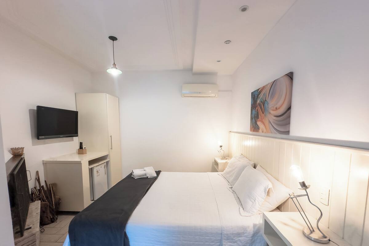 Een hotelkamer met een bed, nachtkastje, lamp, tv aan de muur, airconditioning en een klein kastje.