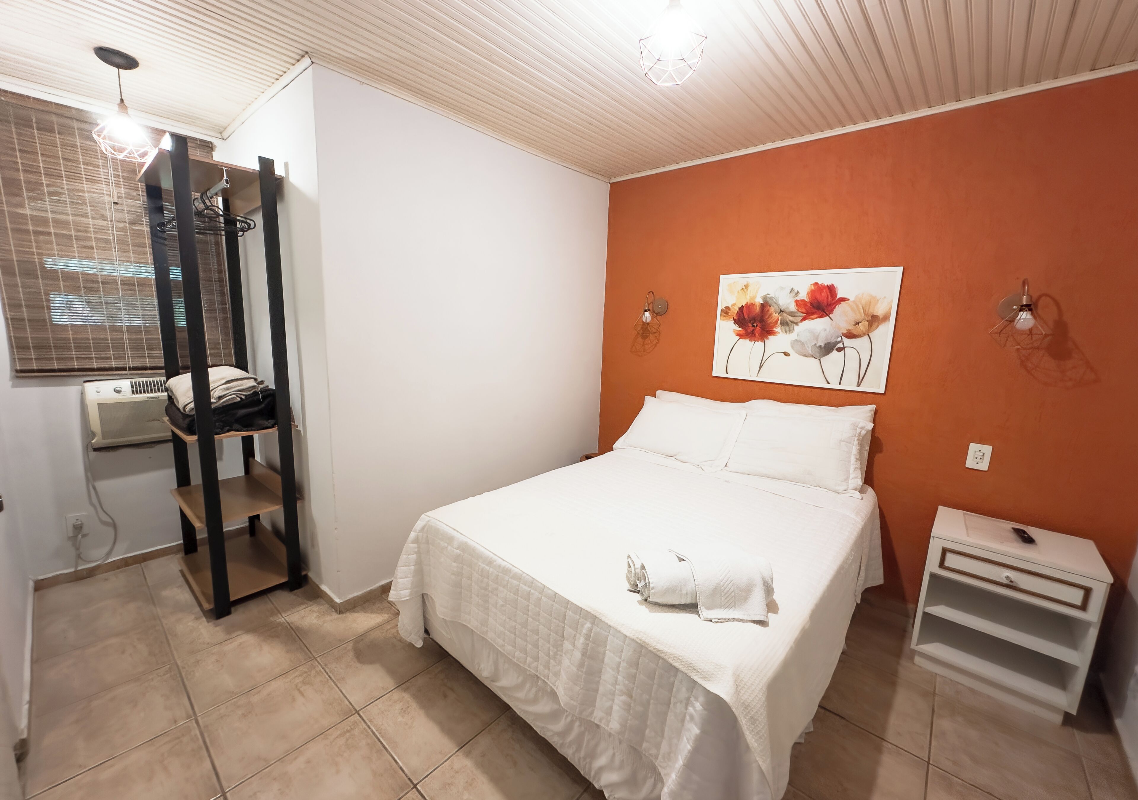 A bedroom with a bed, a nightstand, a wall-mounted shelf, a window with blinds, and a painting.
