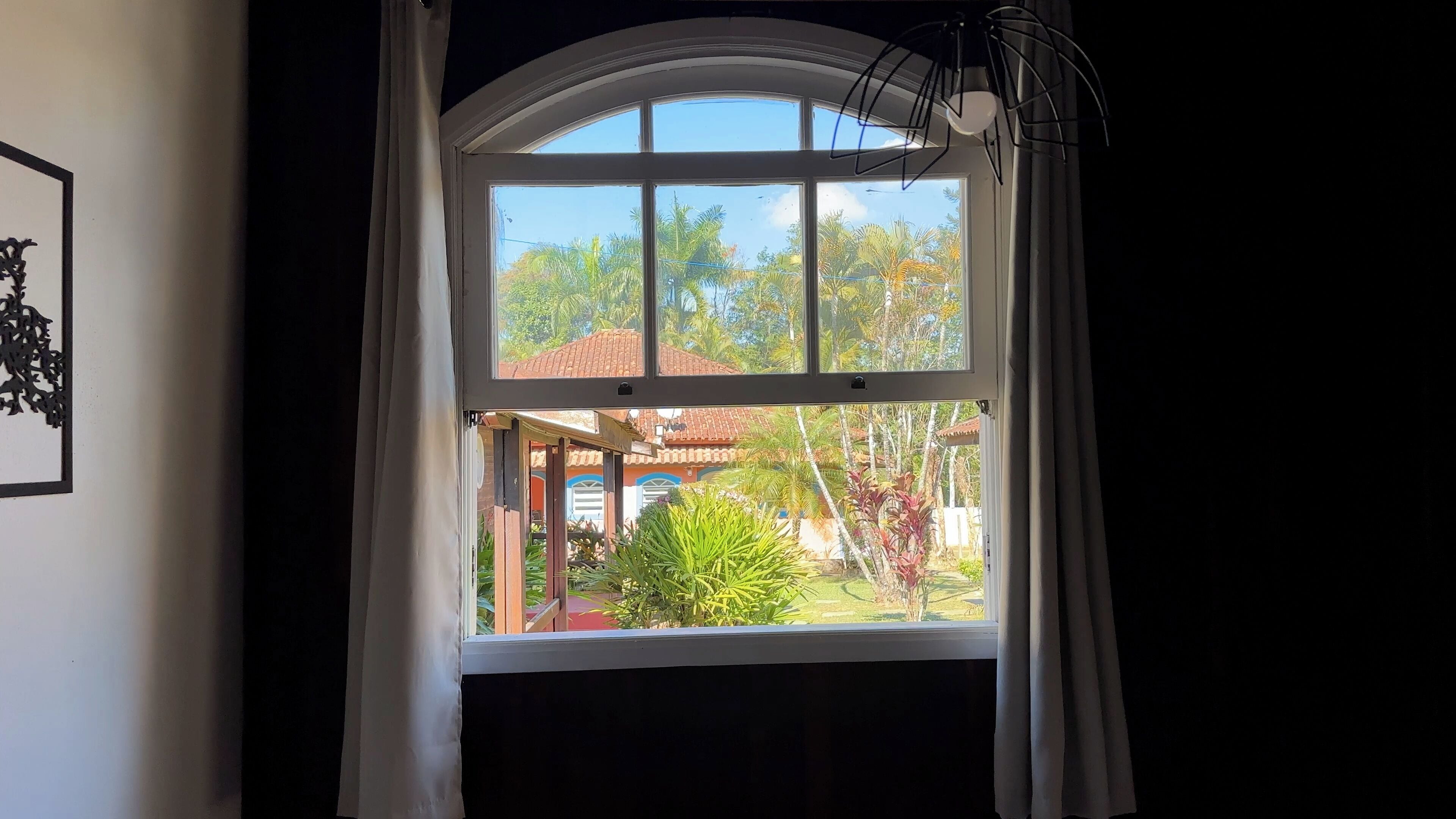 Una ventana con cortinas blancas, vista de una casa con tejado rojo y un jardín con diversas plantas.