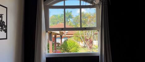 Una ventana con cortinas blancas, vista de una casa con tejado rojo y un jardín con diversas plantas.