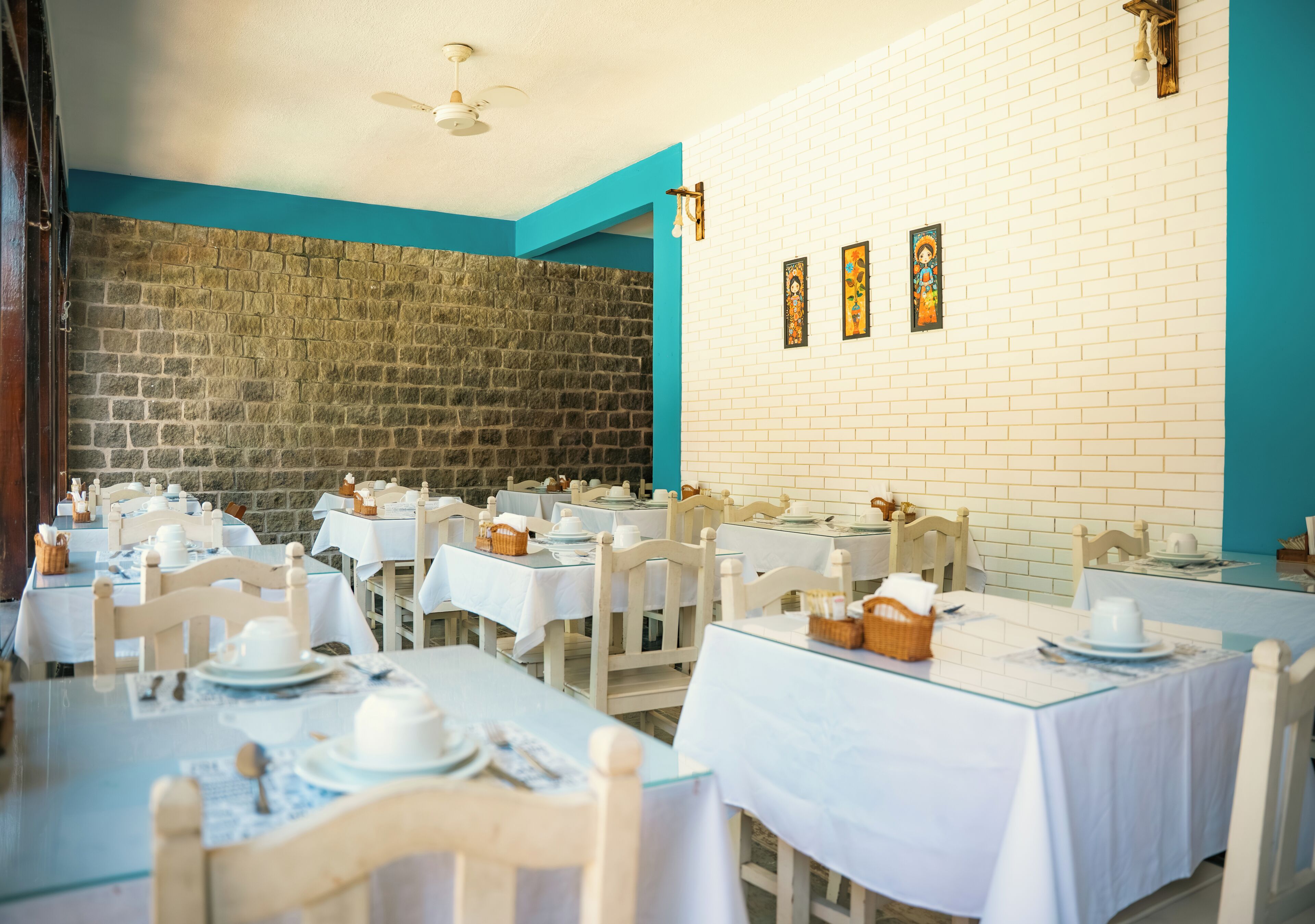 A dining area with tables set for service, a stone wall, and a white brick wall with framed artwork.