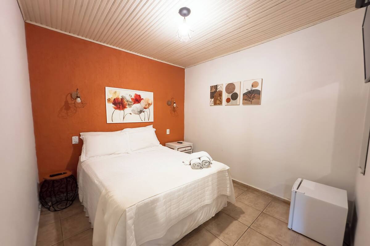 Een slaapkamer met een bed, nachtkastje, een wandlamp, een klein rond tafeltje en een koelkast.