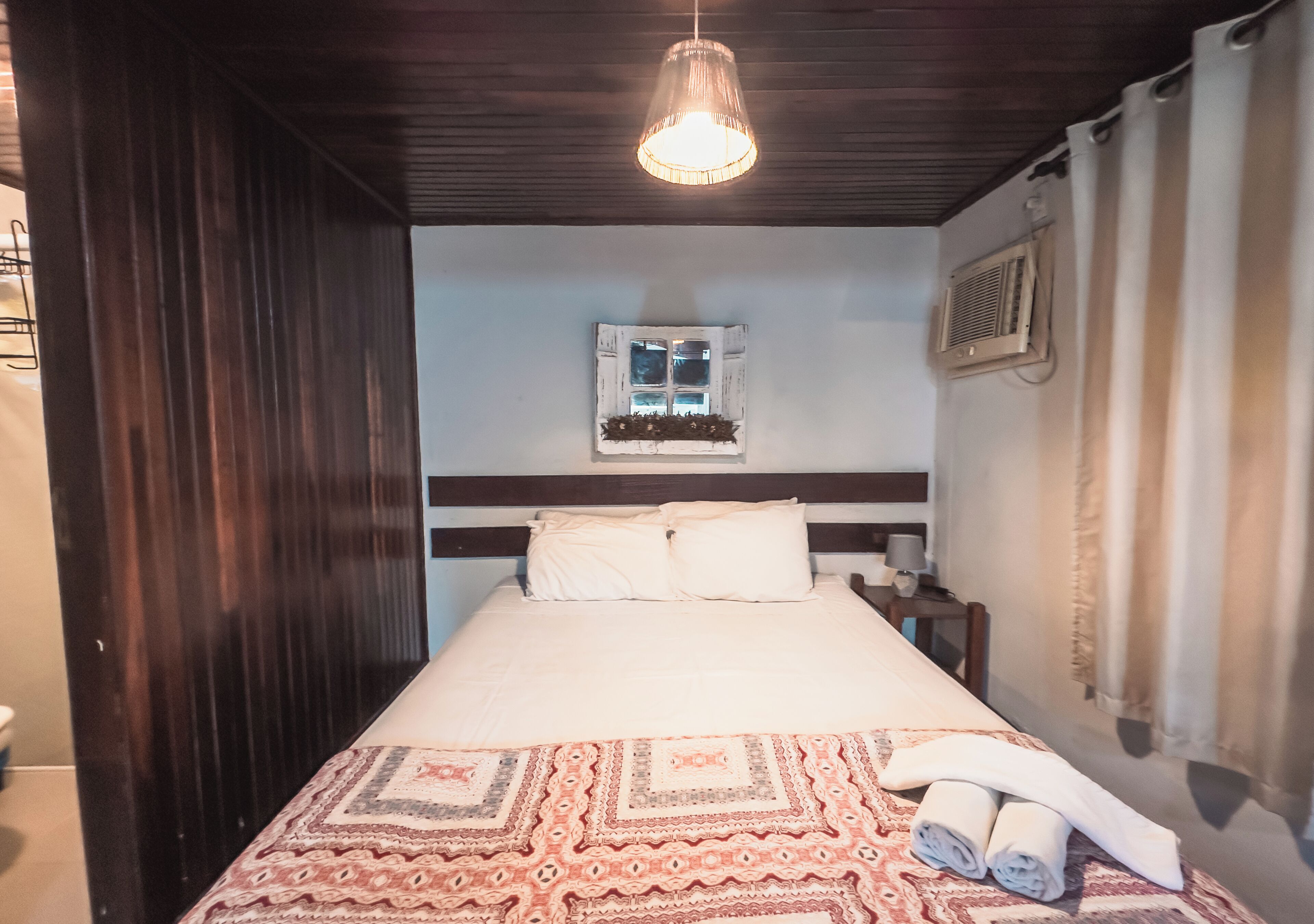 Un dormitorio con una cama, una lámpara colgante, una ventana con contraventanas y un aire acondicionado de pared.