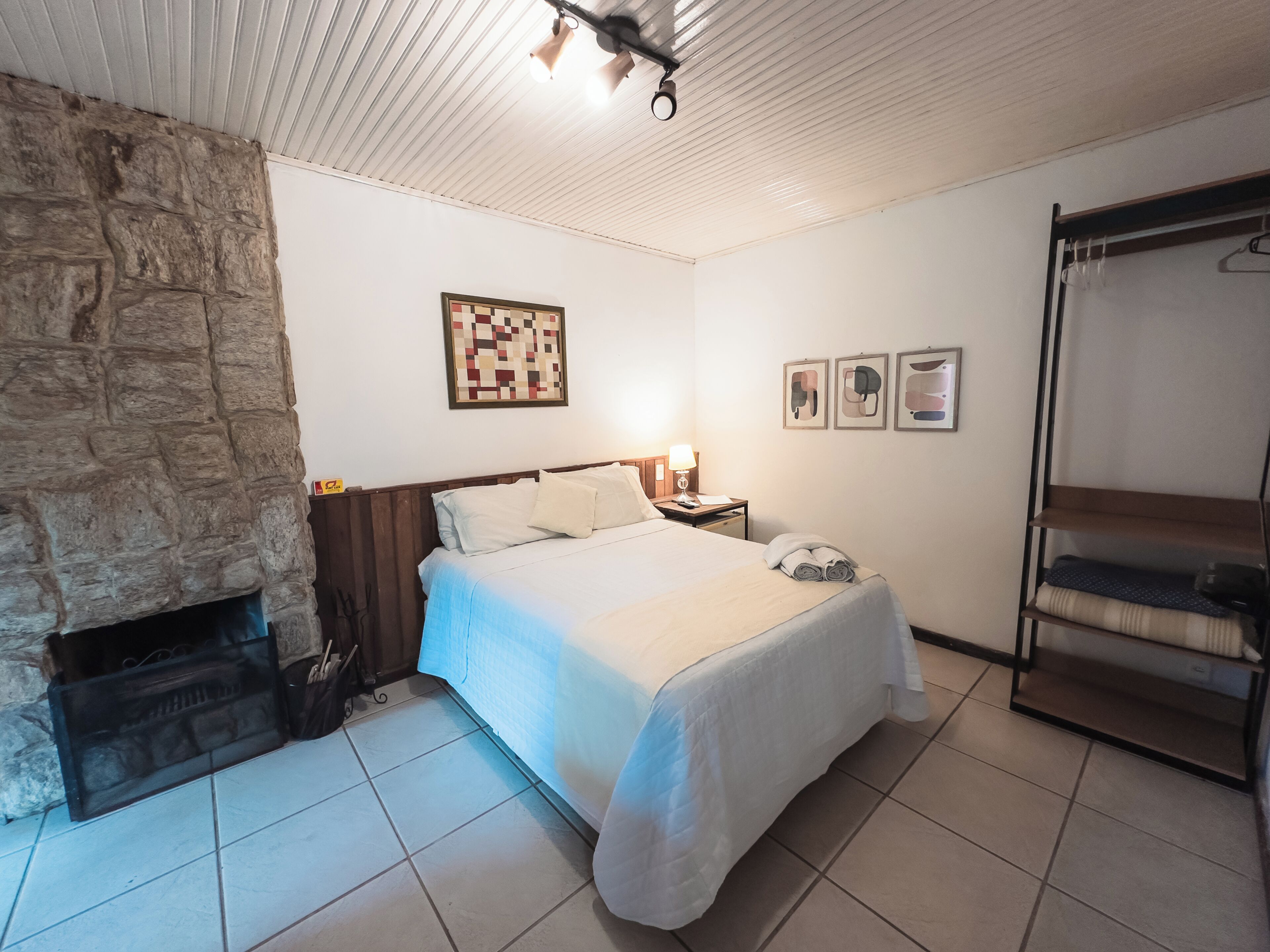 A bedroom with a stone wall, a bed with a headboard, a nightstand, a lamp, a wall-mounted shelf, and a hanging rack.
