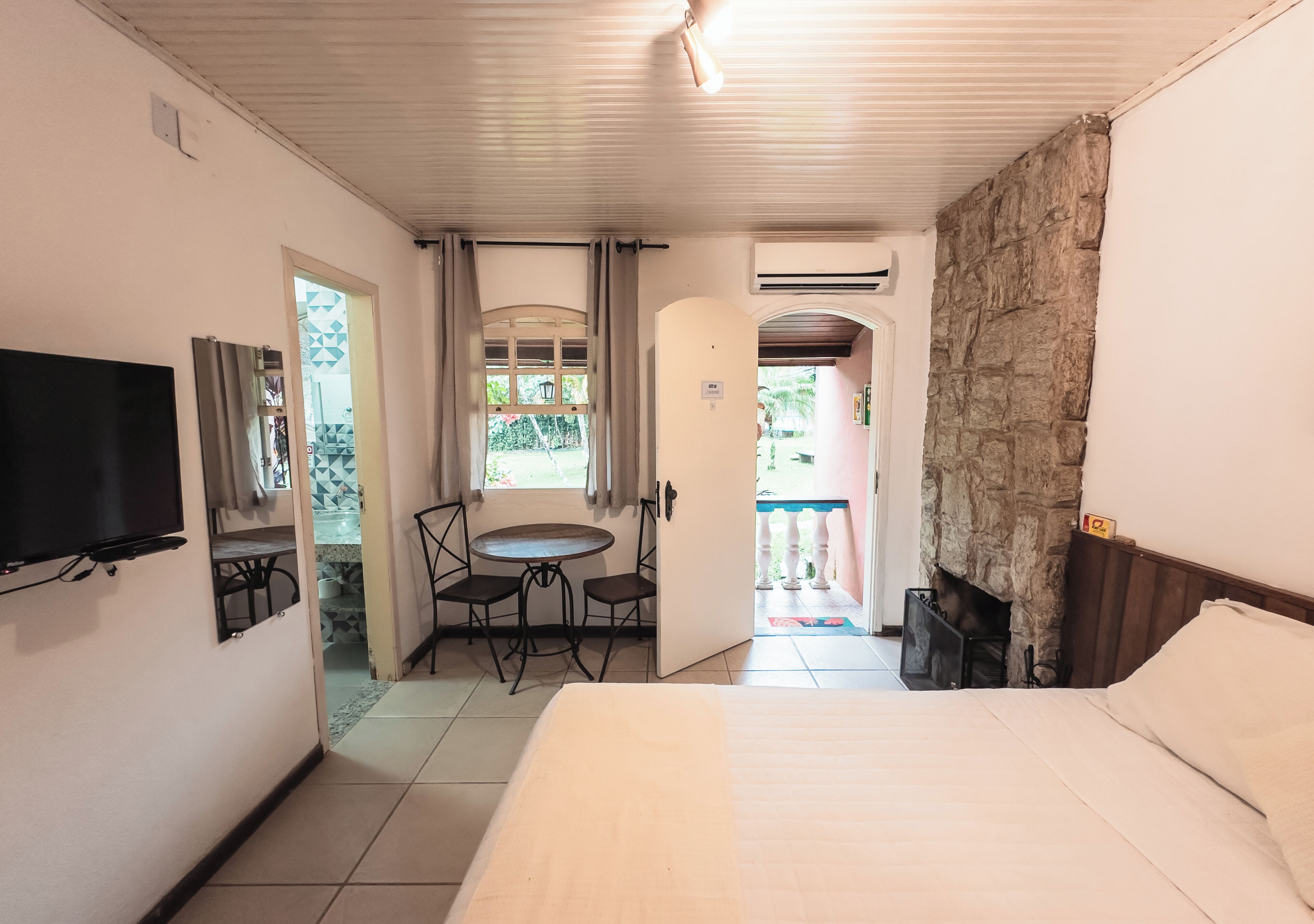 A hotel room with a bed, a TV, a dining table with chairs, a stone wall, and an open door leading to an outdoor area.