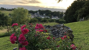 Un jardin orné de rosiers roses, une construction en pierre et une vue lointaine sur des maisons se découpant sur un ciel au coucher du soleil.