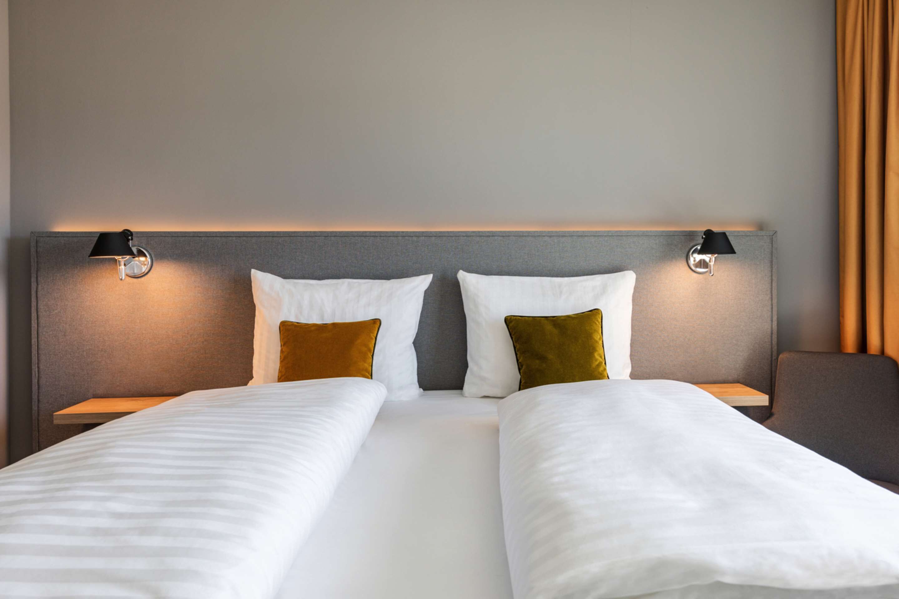 Cama doble con ropa de cama blanca y dos almohadas, una marrón y otra gris, en una habitación de hotel.