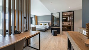 A modern hotel room with a wooden desk, a bed with white linens, a large mirror, and a view of the city. - Piazza Hotel und Restaurant (Aalen)