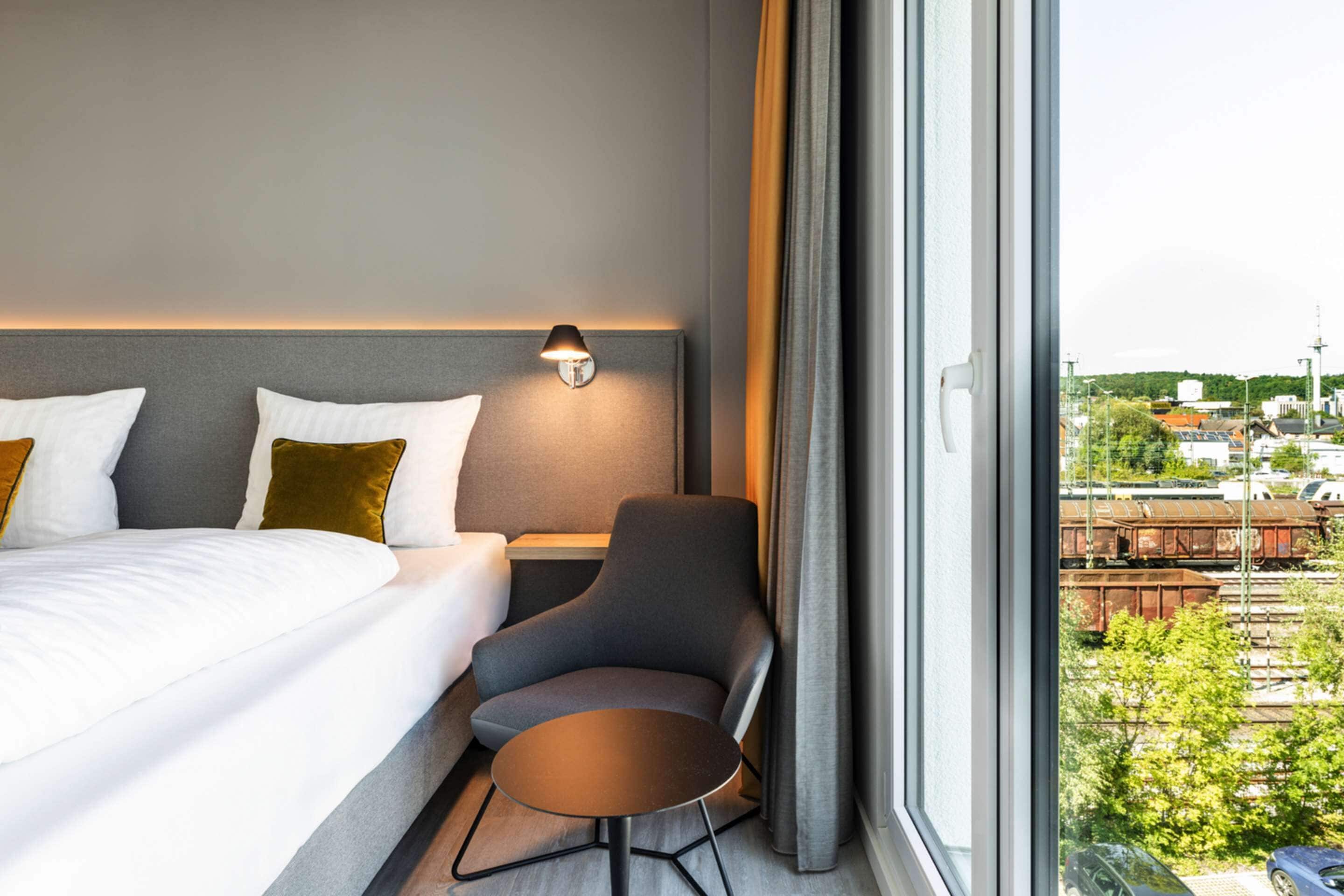 A hotel room with a bed, a desk, a chair, and a view of the city through large windows.