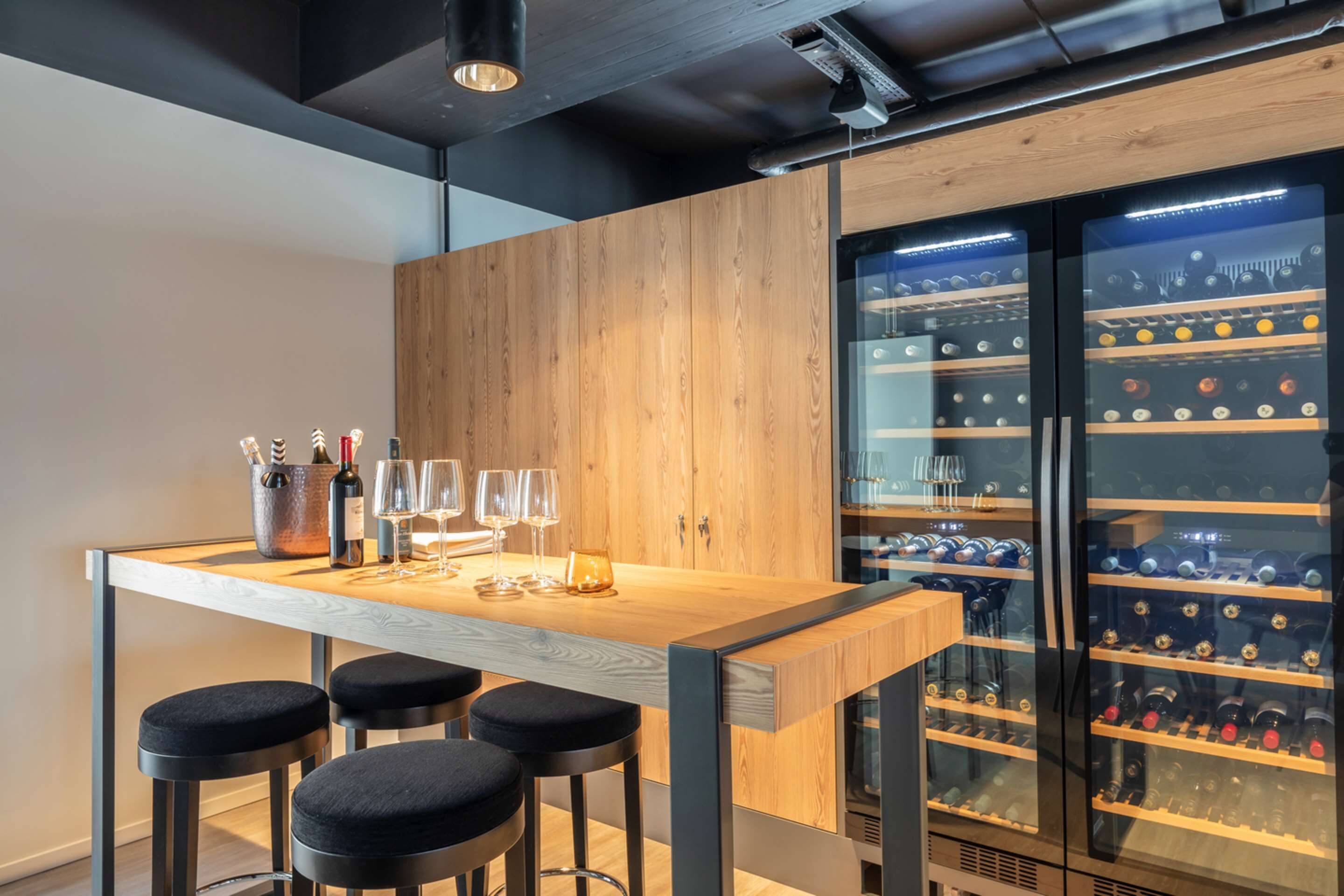 Zona de bar con mostrador de madera, taburetes negros y un enfriador de vinos con botellas.