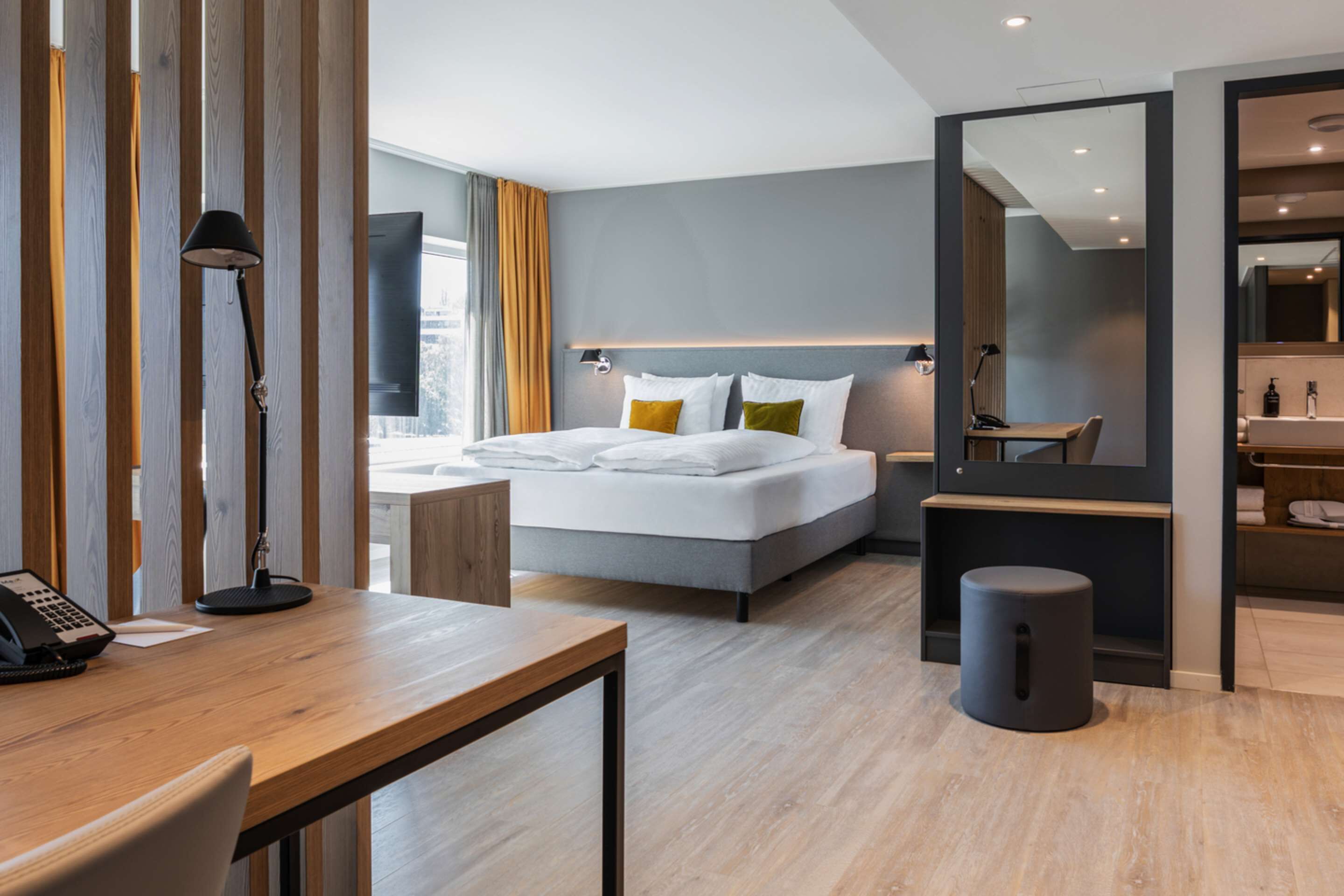 Ett modernt hotellrum med en stor säng, ett skrivbord med en telefon, en spegel och en liten pall.