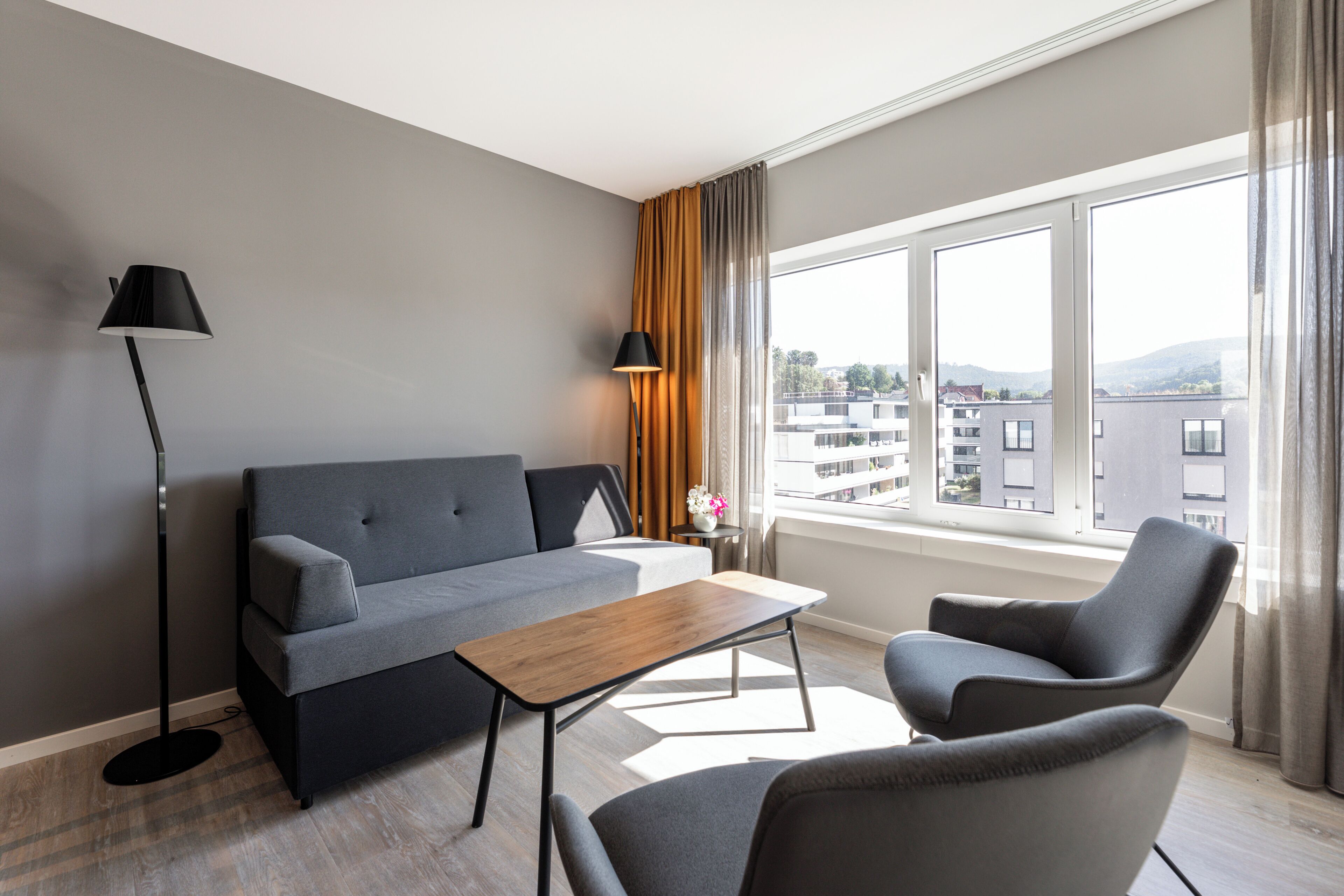 A modern living room with a grey sofa, a wooden coffee table, and two armchairs. There is a floor lamp and a table lamp on a side table. A window with curtains provides a view of buildings outside.