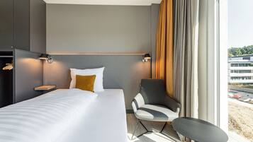 Una habitación de hotel moderna con una cama grande, una silla, una mesita y vistas a un edificio a través de la ventana.