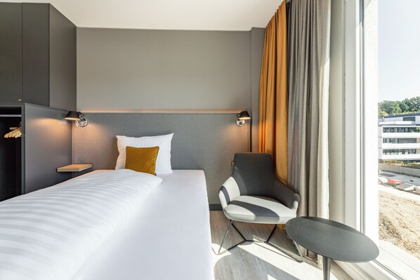 A modern hotel room with a large bed, a chair, a small table, and a view of a building through the window. - Piazza Hotel und Restaurant (Aalen)