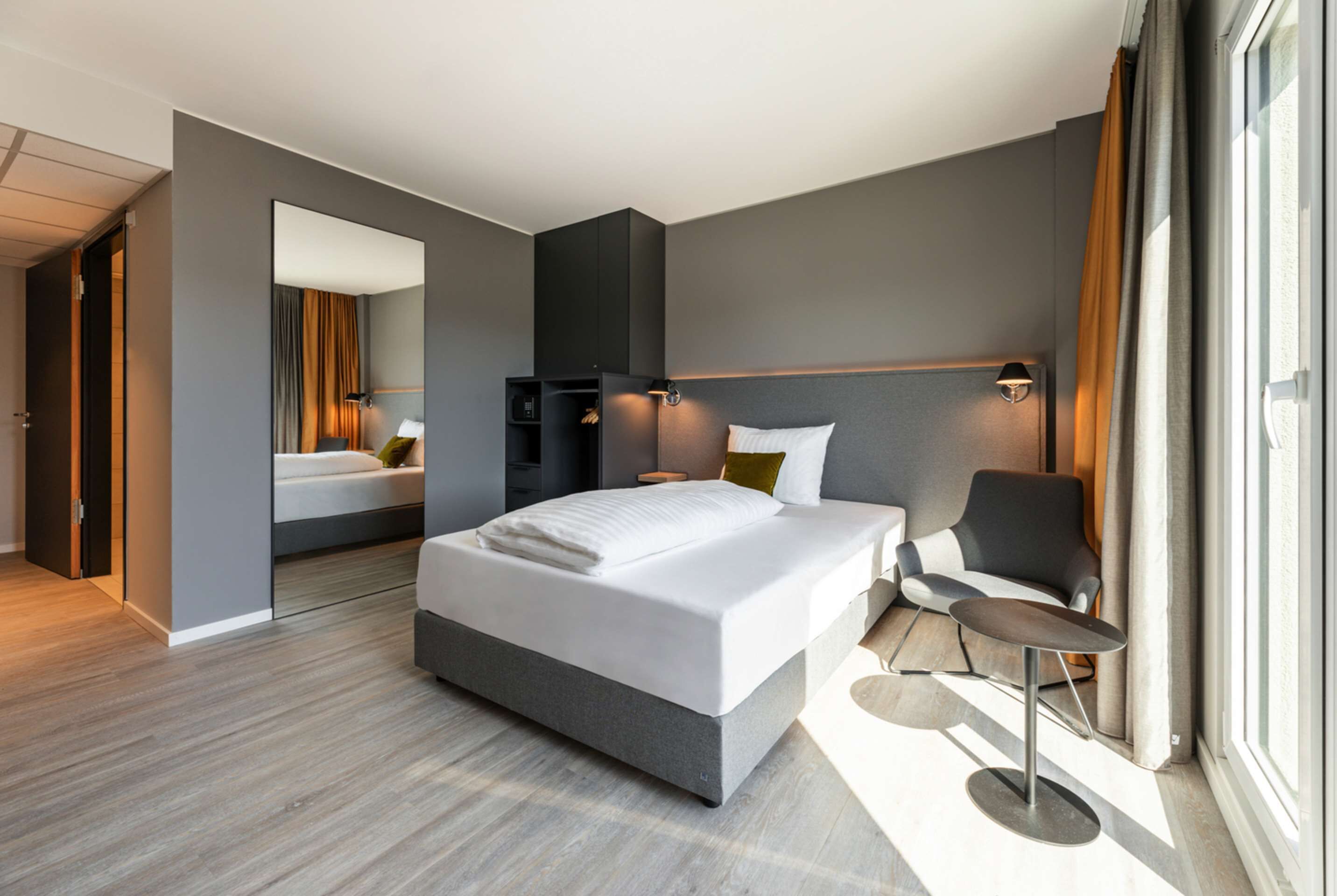A modern hotel room with a large bed, a desk, and a chair. There is a wardrobe and a mirror on one wall, and a window with curtains on the other.