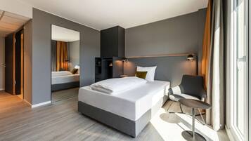 A modern hotel room with a large bed, a desk, and a chair. There is a wardrobe and a mirror on one wall, and a window with curtains on the other.