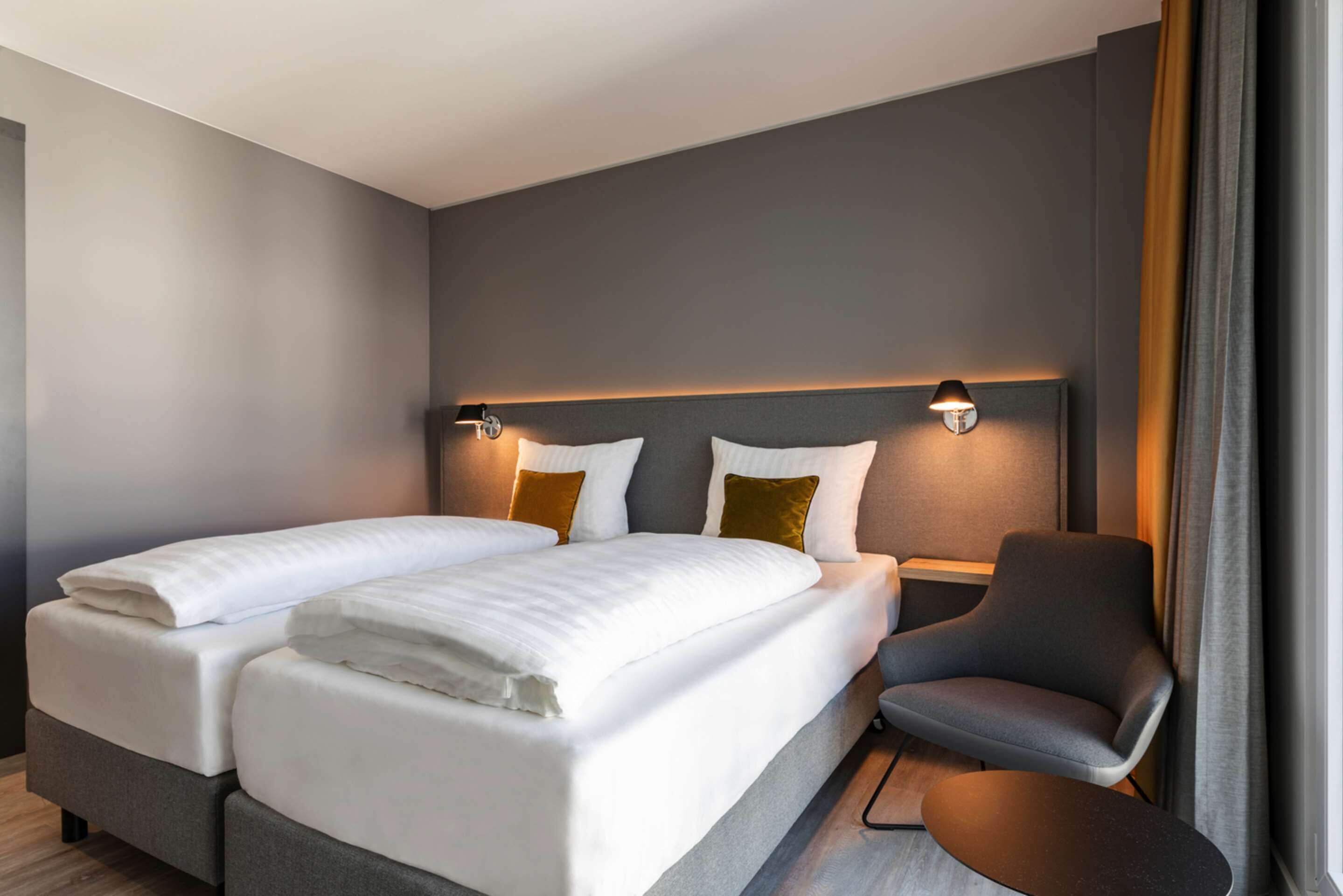 A modern hotel room with a double bed, two bedside tables, a chair, and a large window with curtains.