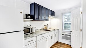 A kitchen with white cabinets, a black range hood, a microwave, a coffee maker, and a window with blinds. - Ocean Breeze Motel - Kitchenette (South Yarmouth)