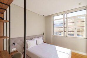 A bedroom with a bed, a chair, a shelf, and a window with a view of buildings.