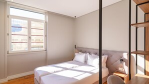 Quarto com cama, mesa de cabeceira, cadeira e janela com vista para edifícios.