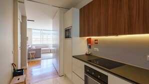Cozinha moderna com micro-ondas, forno e armários de madeira. Um pequeno banquinho de madeira e uma lixeira são visíveis.
