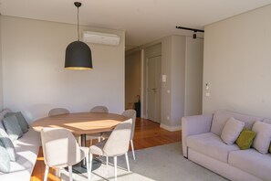 A modern living room with a round wooden table, white chairs, a grey sofa, and a black pendant light.
