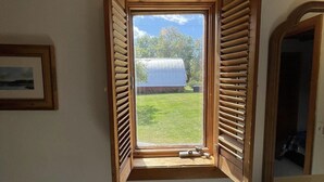 Una habitación con una pequeña ventana con contraventanas de madera, una pared blanca, un cuadro enmarcado, un dispensador de toallas blanco y un espejo.