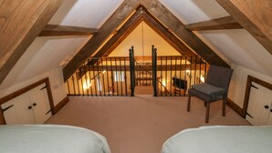 A cozy attic bedroom with a sloped ceiling, wooden beams, a bed, a chair, and a view of the lower level. - The Old Dairy (Clarbeston Road)