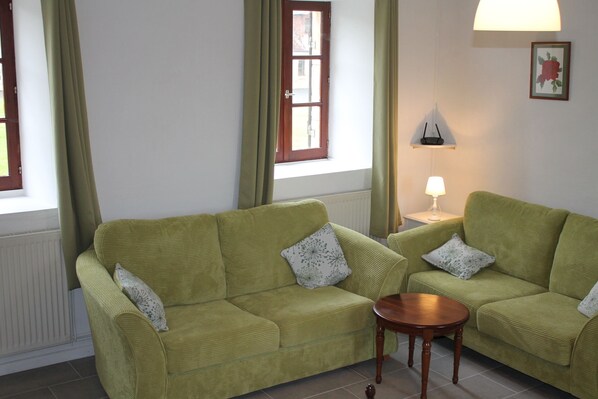 A living room with two green sofas, a small wooden table, a lamp, and a window with curtains.