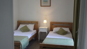 A room with two wooden beds, each with a green pillow and white bedding. There is a small bedside table with a lamp between the beds.