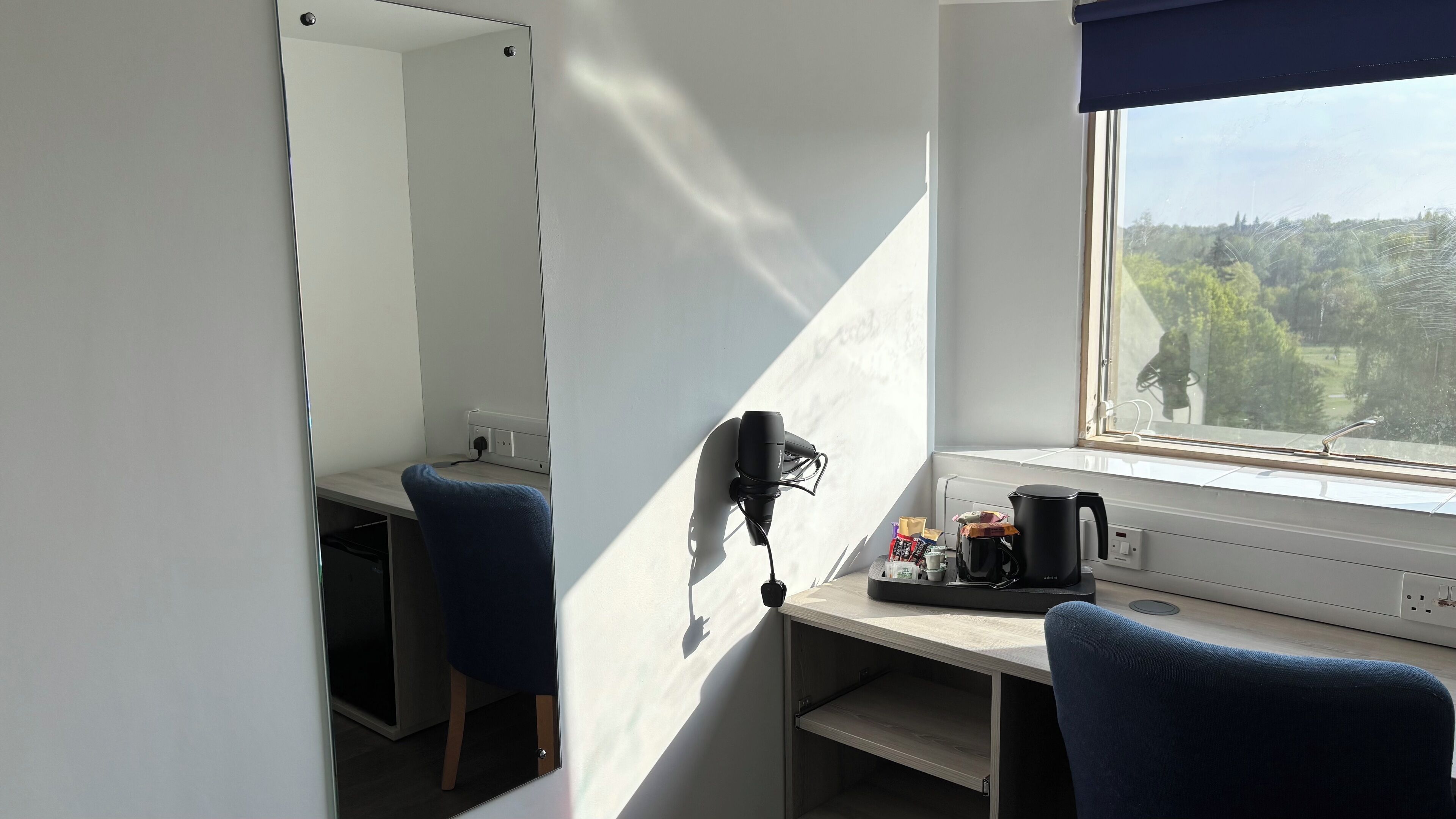 A room with a desk, a chair, a mirror, a window with a view of trees, and a coffee maker.