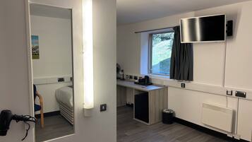 Une chambre d’hôtel de taille réduite, comprenant un lit, un bureau, un miroir, une fenêtre avec des rideaux, un téléviseur fixé au mur et un radiateur.