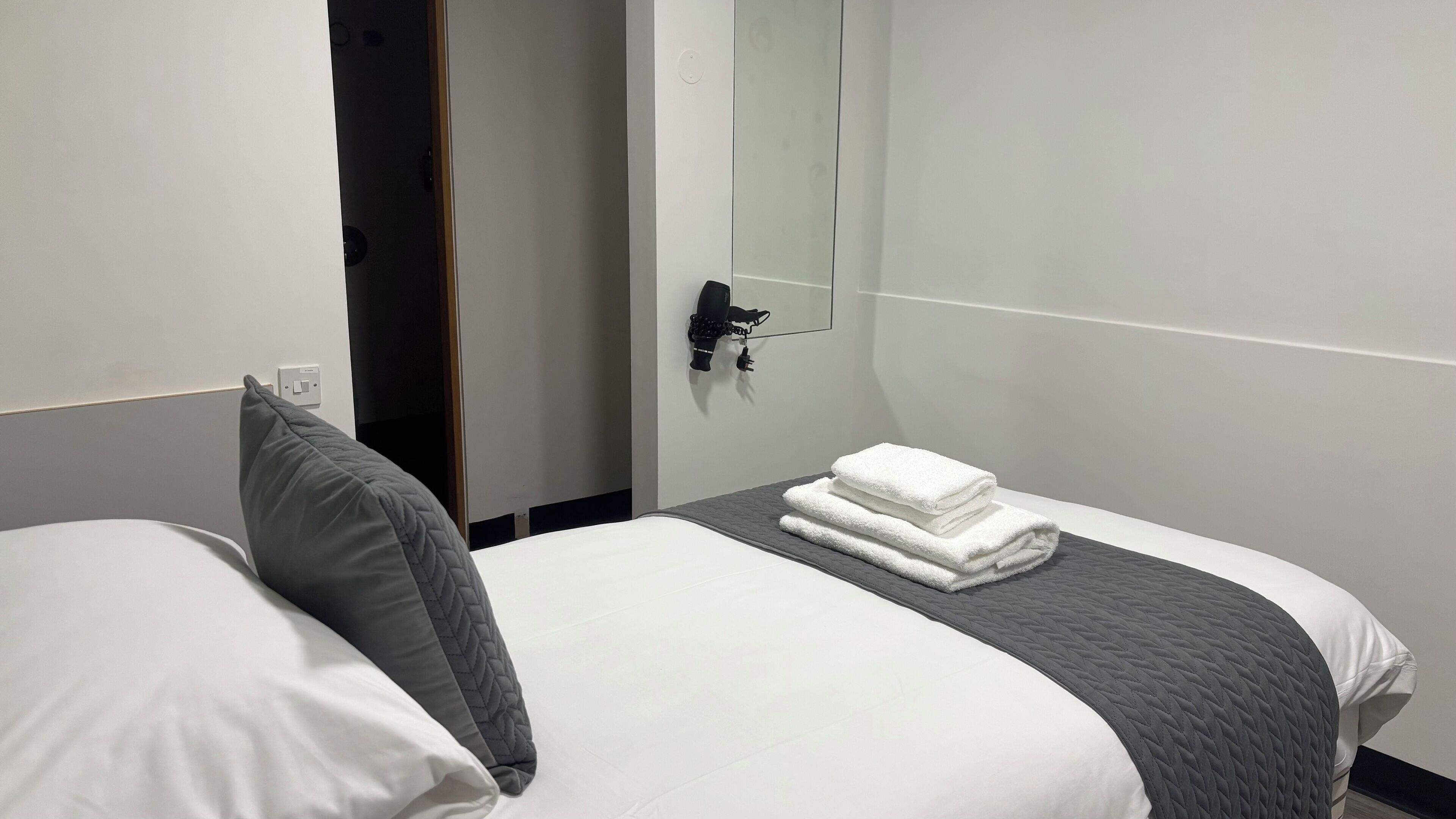 A hotel room with a bed, a grey quilted pillow, a folded white towel, and a hairdryer on a wall-mounted stand.