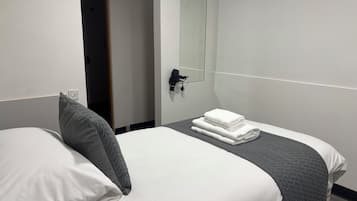 A hotel room with a bed, a grey quilted pillow, a folded white towel, and a hairdryer on a wall-mounted stand.