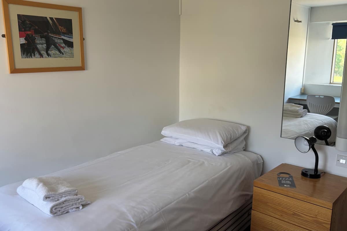 A neatly made bed with white linens and a folded towel, a wooden nightstand with a lamp, a framed picture on the wall, and a mirror reflecting the room.