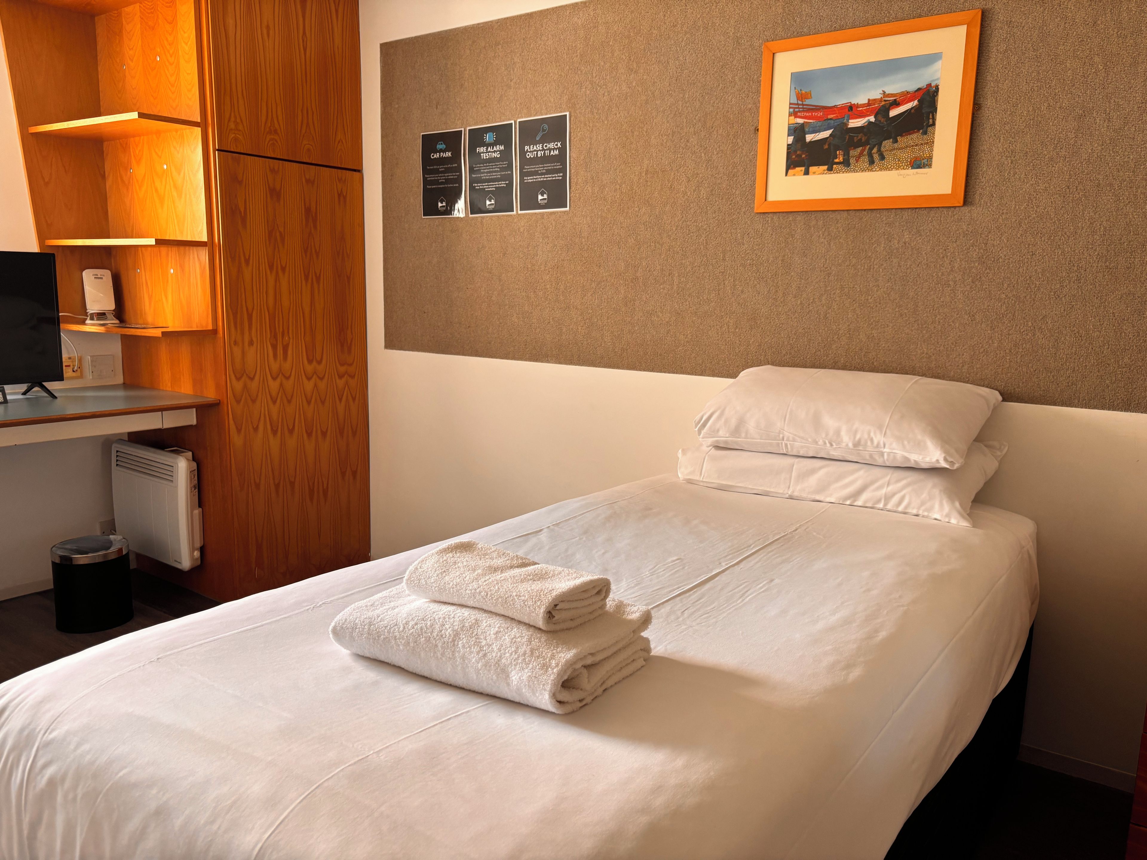 Una habitación de hotel con una cama, un escritorio con una computadora, un radiador y un cuadro en la pared.