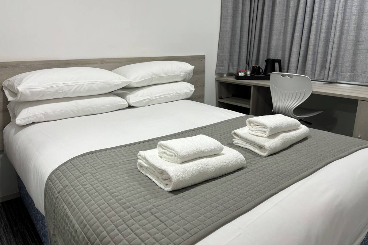 A neatly made bed with white linens and a grey quilted bedspread. There are two folded towels on the bed.