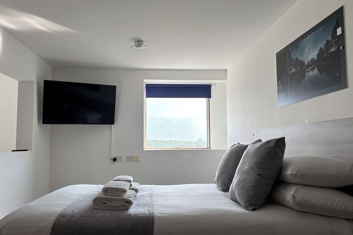 A bedroom with a bed, a flat-screen TV mounted on the wall, a window with a view, and a painting above the bed.