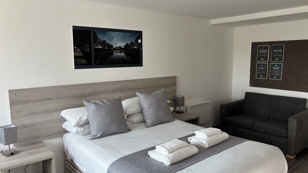 A modern hotel room with a large bed, grey and white bedding, a grey armchair, and a wall-mounted picture. - Broadview Hotel (Norwich)