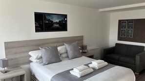 A modern hotel room with a large bed, grey and white bedding, a grey armchair, and a wall-mounted picture. - Broadview Hotel (Norwich)