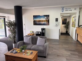 A modern hotel lobby with a grey sofa, a wooden table with magazines, a large window, a black pillar, a recycling bin, and a sign indicating room directions.