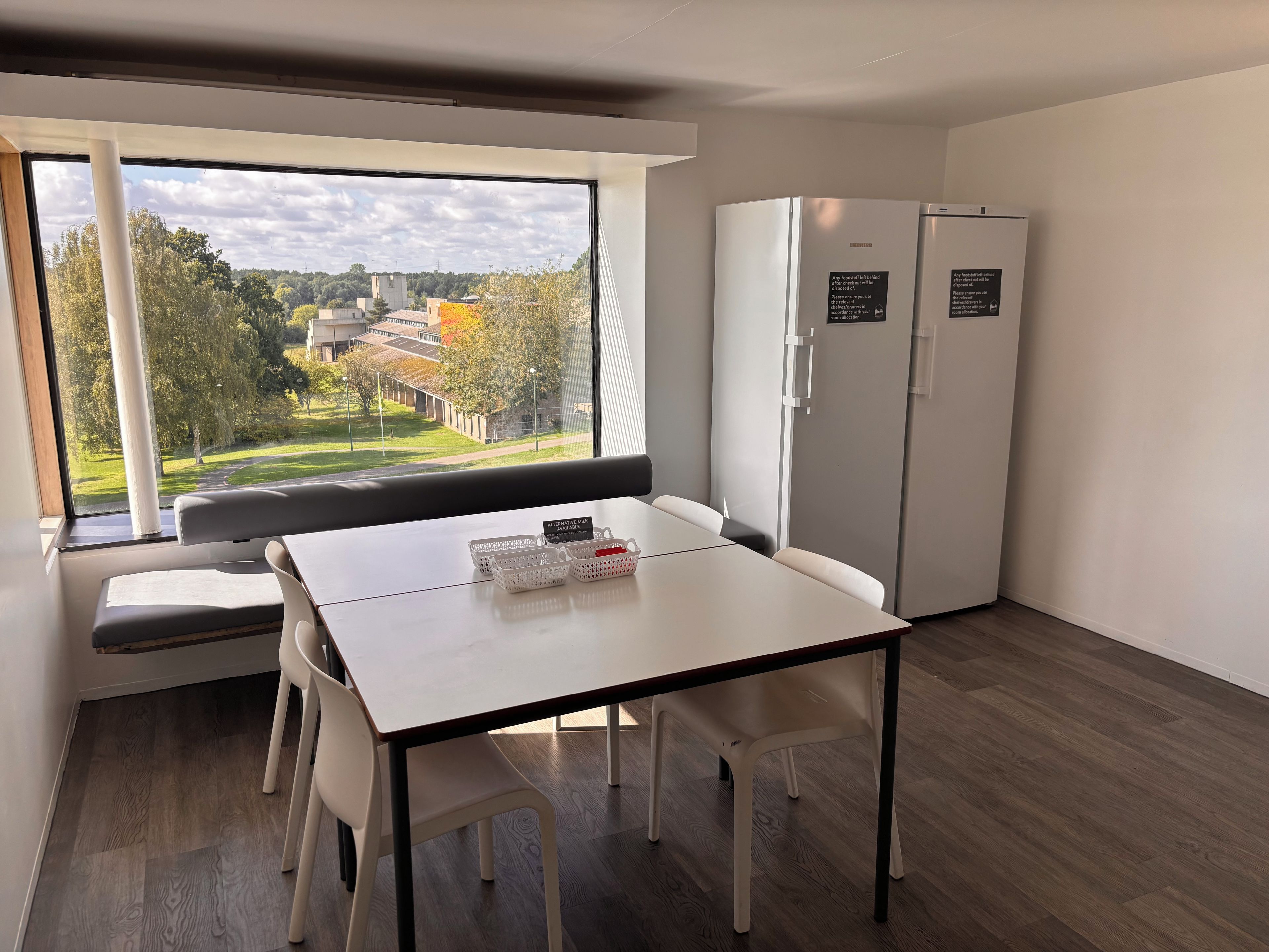 A modern room with a large window, a white table, two white chairs, a bench, and a white refrigerator.