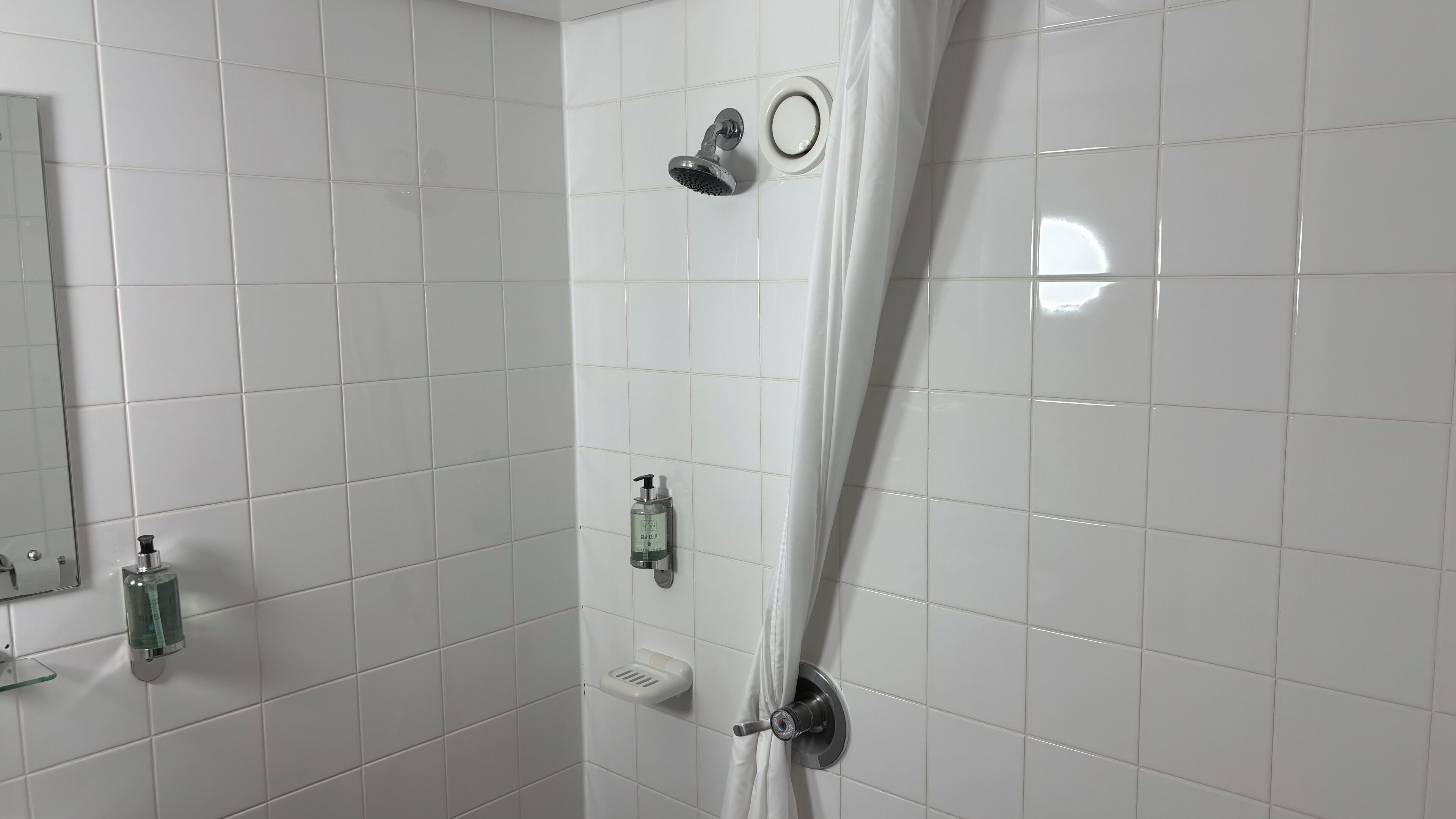 Un baño moderno con azulejos blancos, ducha con cabezal de ducha de mano, dispensador de jabón y un plato de cristal para jabón.